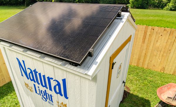 An elevated, close-up shot of the white Natural Light branded shed, highlighting a solar panel mounted across its sloped roof in a grassy backyard.