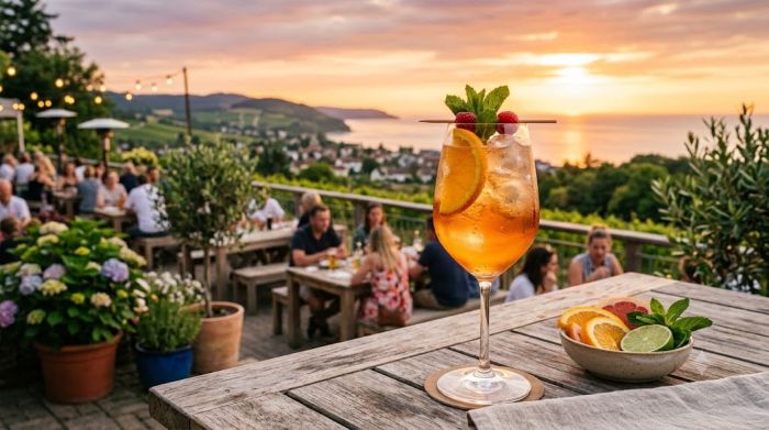 An IPA cocktail in a tulip glass on a wooden table overlooks a crowded outdoor patio at sunset.