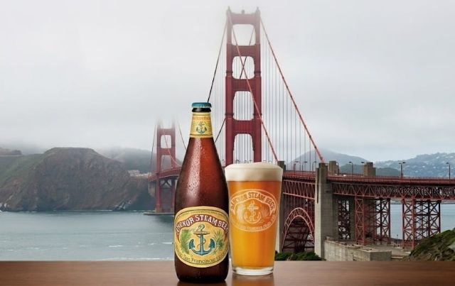 A bottle and a glass of Anchor Steam beer sit on a wooden table, with the Golden Gate Bridge wrapped in fog in the background.