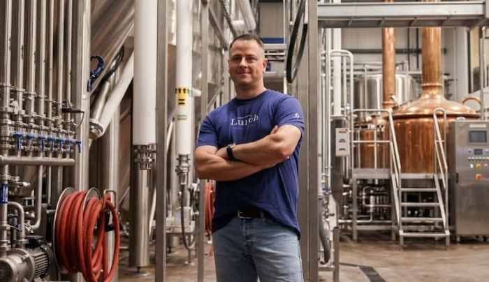 Dan Kleban with folded arms standing inside a modern brewery with stainless steel and copper equipment in background.