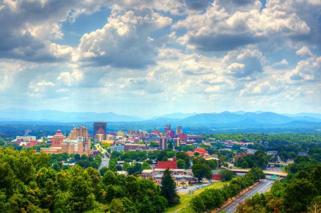 An elevated, wide-angle shot of the Asheville, North Carolina skyline nestled against the Blue Ridge Mountains under a partly cloudy sky.