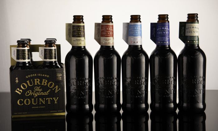 A row of Goose Island Bourbon County Brand Stout bottles and a 4-pack carton against a plain backdrop.