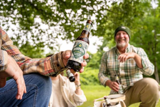 A close-up shot of a person's hand holding a bottle of Harpoon "Flannel Friday" beer, with other people wearing plaid shirts sitting in a grassy, wooded outdoor area blurred in the background.
