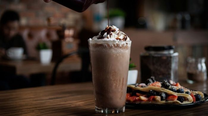 Chocolate syrup being drizzled over whipped cream on top of a chocolate Stout beer float in a tall glass, with a plate of berry crepes nearby on a wooden table.