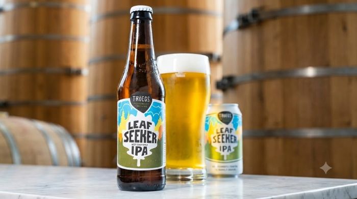 A bottle, glass, and can of Tröegs Leaf Seeker IPA on a counter in front of wooden barrels.