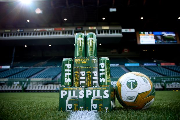 A stacked pyramid of green Widmer Brothers PILS beer cans and boxes stands next to a Portland Timbers soccer ball on stadium grass.