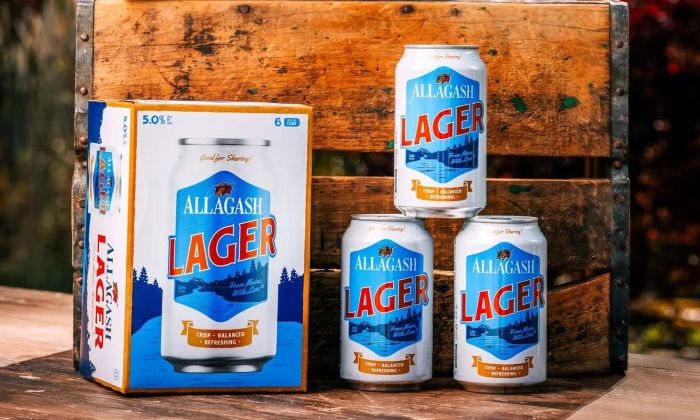 A stacked tower of three Allagash Lager beer cans stands next to a matching six-pack cardboard box, all set against a rustic, weathered wooden background.