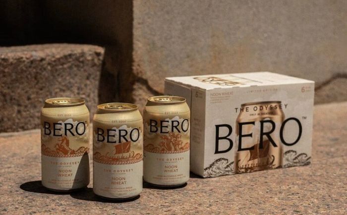 Three “The Odyssey” themed non-alcoholic beer cans and a six-pack box labeled "BERO" and "Noon Wheat" on a concrete step.
