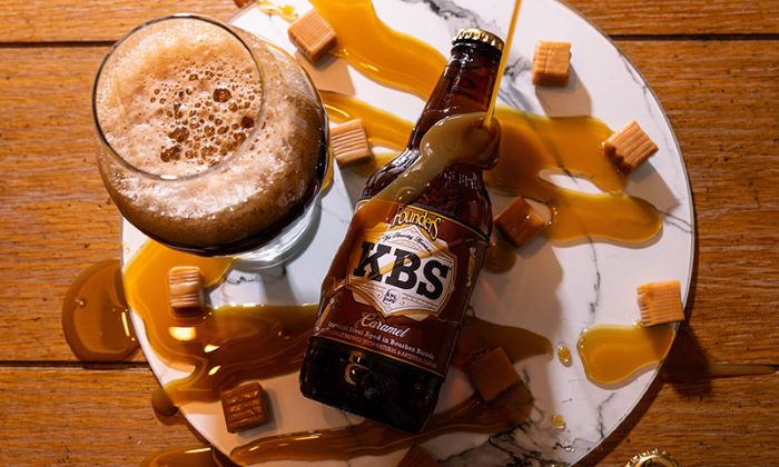 An overhead shot of a bottle of Founders KBS Caramel stout lying on a marble platter, surrounded by spilled caramel sauce, loose caramel cubes, and a foamy glass of dark beer.