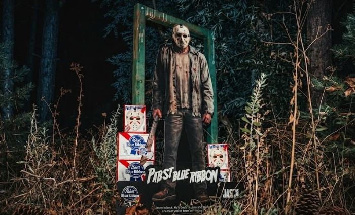 A life-sized Jason Voorhees cardboard cutout holding a blood-splattered axe stands outdoors at night, surrounded by Pabst Blue Ribbon beer boxes featuring his iconic hockey mask design