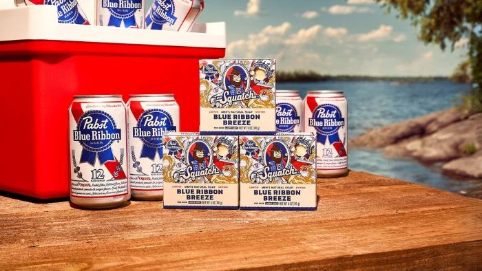 Dr. Squatch Blue Ribbon Breeze bar soaps and Pabst Blue Ribbon beer cans are arranged on a wooden table outdoors next to a red cooler and a scenic lake.