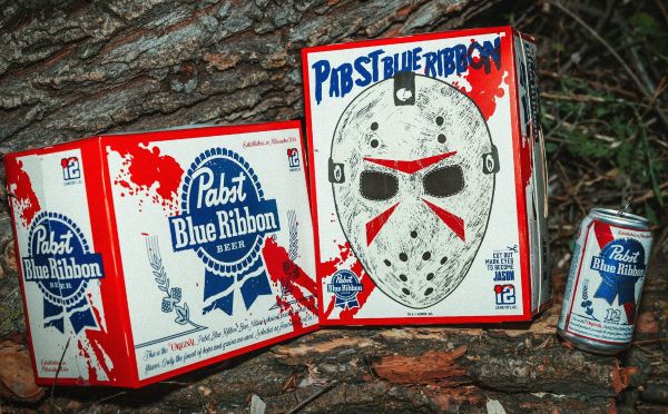 Two boxes and a single can of Pabst Blue Ribbon beer sitting outdoors on a wooden surface, featuring the limited-edition horror-themed Jason Universe collaboration packaging with a large hockey mask printed on the front box.