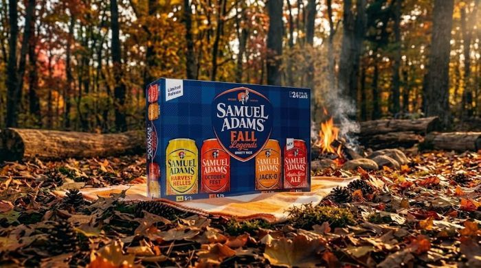 A 24-can variety pack of Samuel Adams "Fall Legends" beer sits on a plaid blanket in a sunlit autumn forest, surrounded by fallen leaves with a small campfire burning softly in the background.
