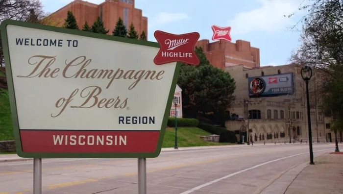 A large green and white sign on a city street reading "Welcome to The Champagne of Beers Region Wisconsin" with a Miller High Life logo, set against a background of large brick brewery buildings.