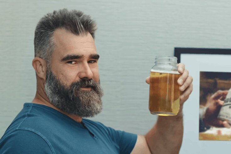 Former NFL player Jason Kelce smiles at the camera while holding up a mason jar filled with his urine. 