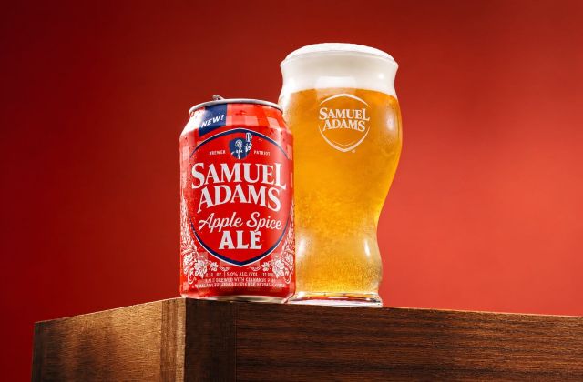 Samuel Adams Apple Spice Ale can beside a glass of golden beer against a red backdrop.