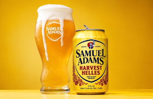 Samuel Adams Harvest Helles can beside a glass of golden lager topped with a thick white foam head.