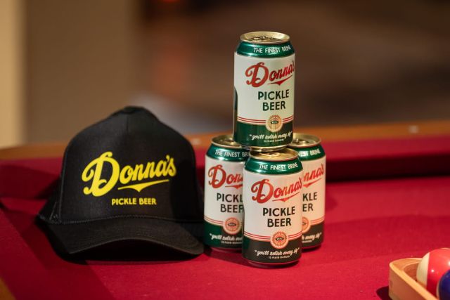 Donna’s Pickle Beer cans stacked beside a black branded cap on a red pool table.
