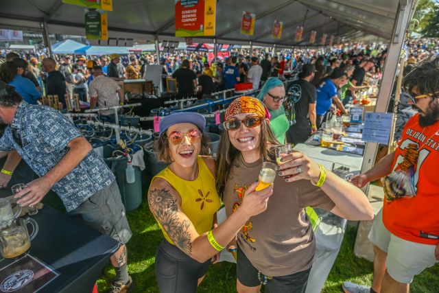 Two festivalgoers raise glasses of beer at the 2026 Great American Beer Festival in Denver, Colorado, surrounded by crowds enjoying the outdoor event.