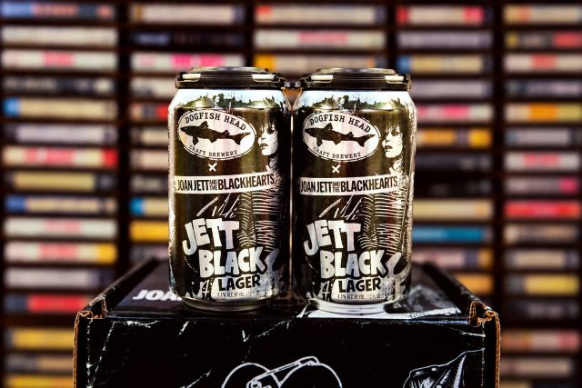 Two cans of Dogfish Head and Joan Jett’s Jett Black Lager on a collectible box in front of shelves of cassette tapes.