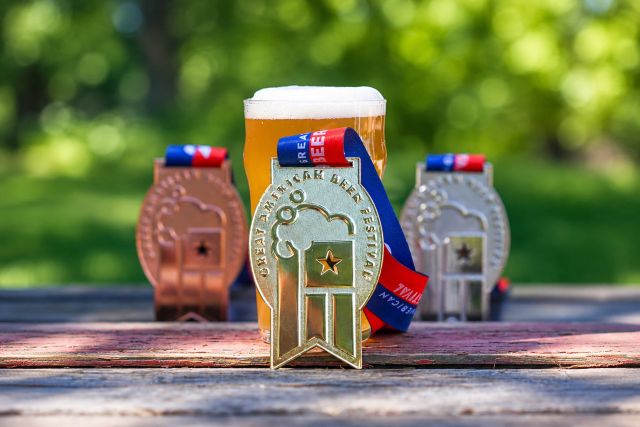 Gold, silver and bronze Great American Beer Festival medals displayed with a glass of amber beer on an outdoor picnic table.